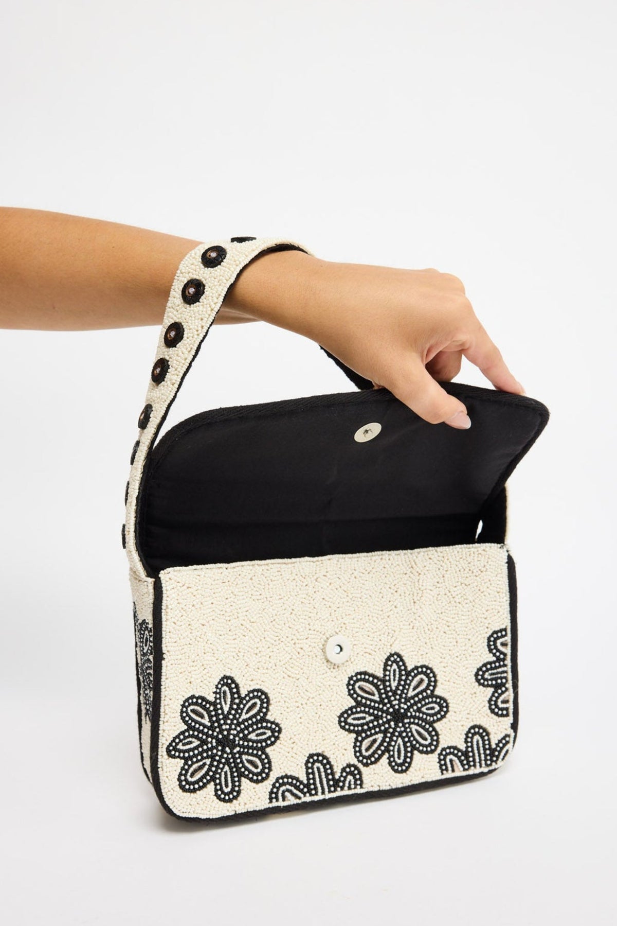 A hand holding a small rectangular shoulder bag with a black interior and a cream exterior decorated with black floral embroidery and a matching embroidered strap.