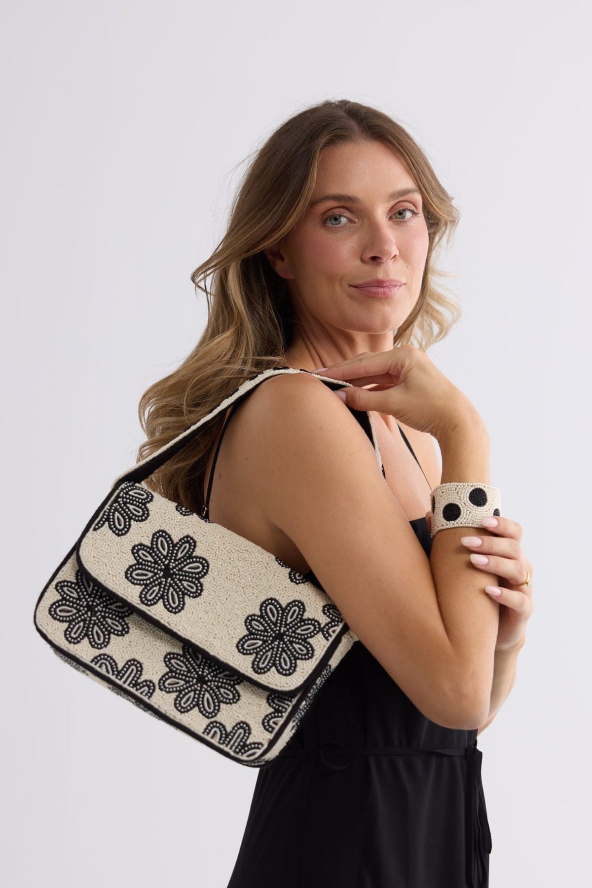 A woman wearing a black dress carries a beige shoulder bag with black floral embroidery against a plain light gray studio background.