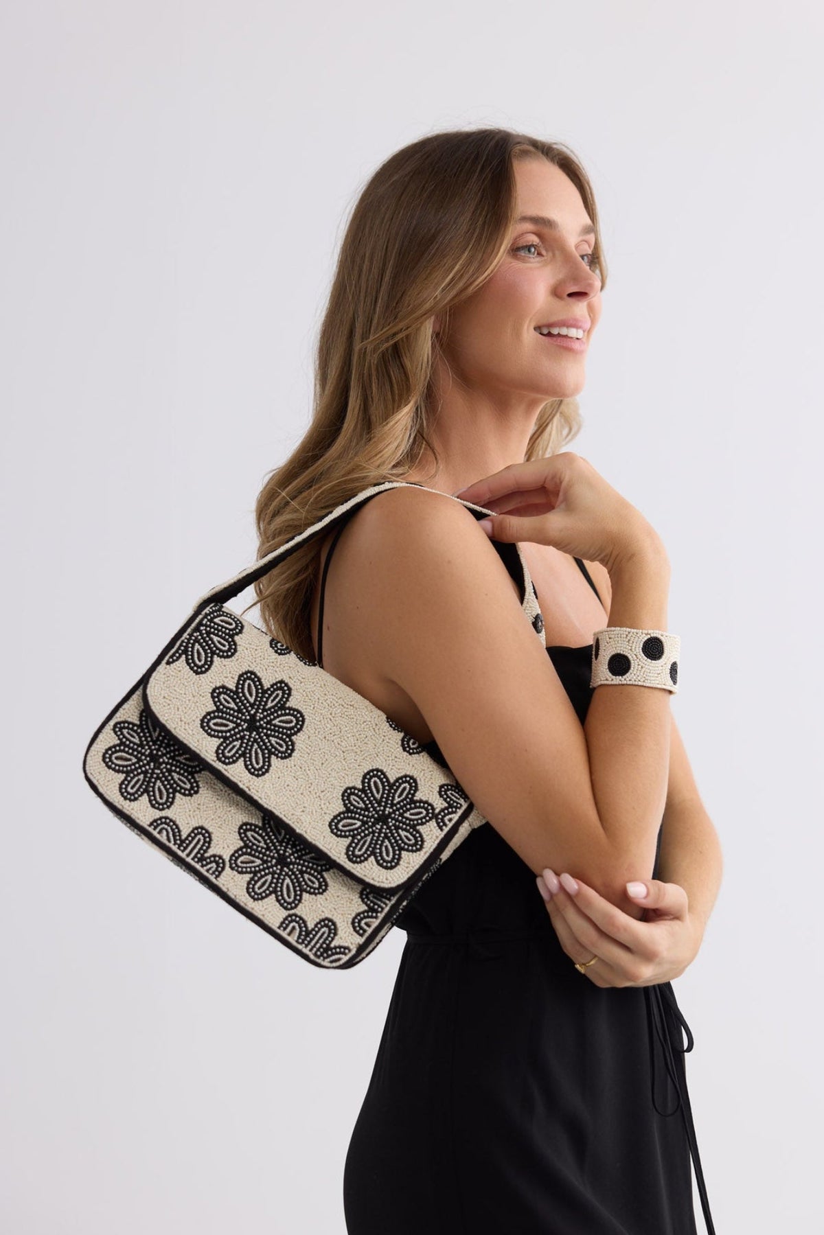 A woman in a black dress carries a black and beige floral patterned shoulder bag against her shoulder, posing against a light gray studio background.