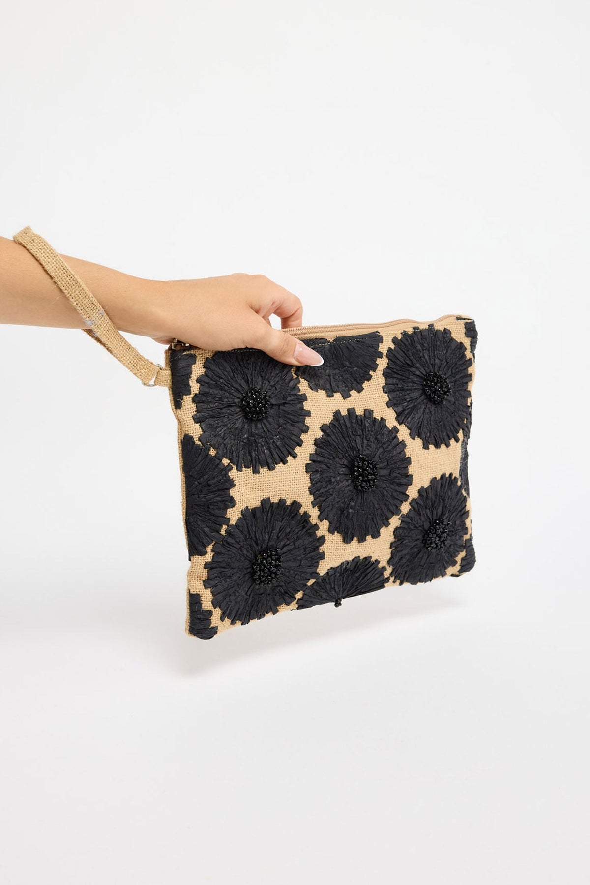 Rectangular zip clutch in natural woven raffia with bold black 3D raffia flower appliqués covering the front panel, held by hand against a white background