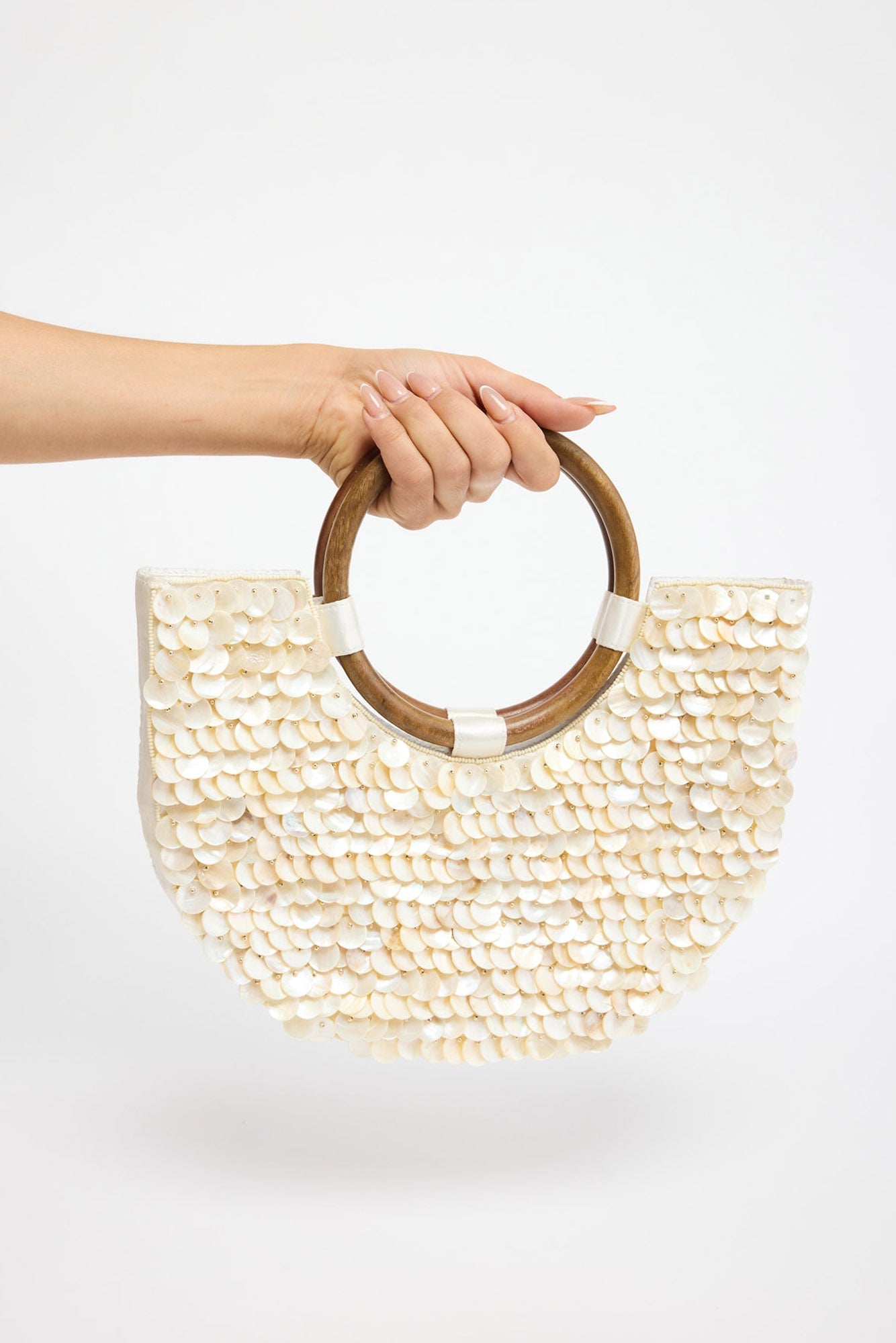 Half-moon shaped bag fully covered in overlapping cream shell discs with a warm iridescent sheen, held by a round tortoiseshell-tone acrylic handle against a white background