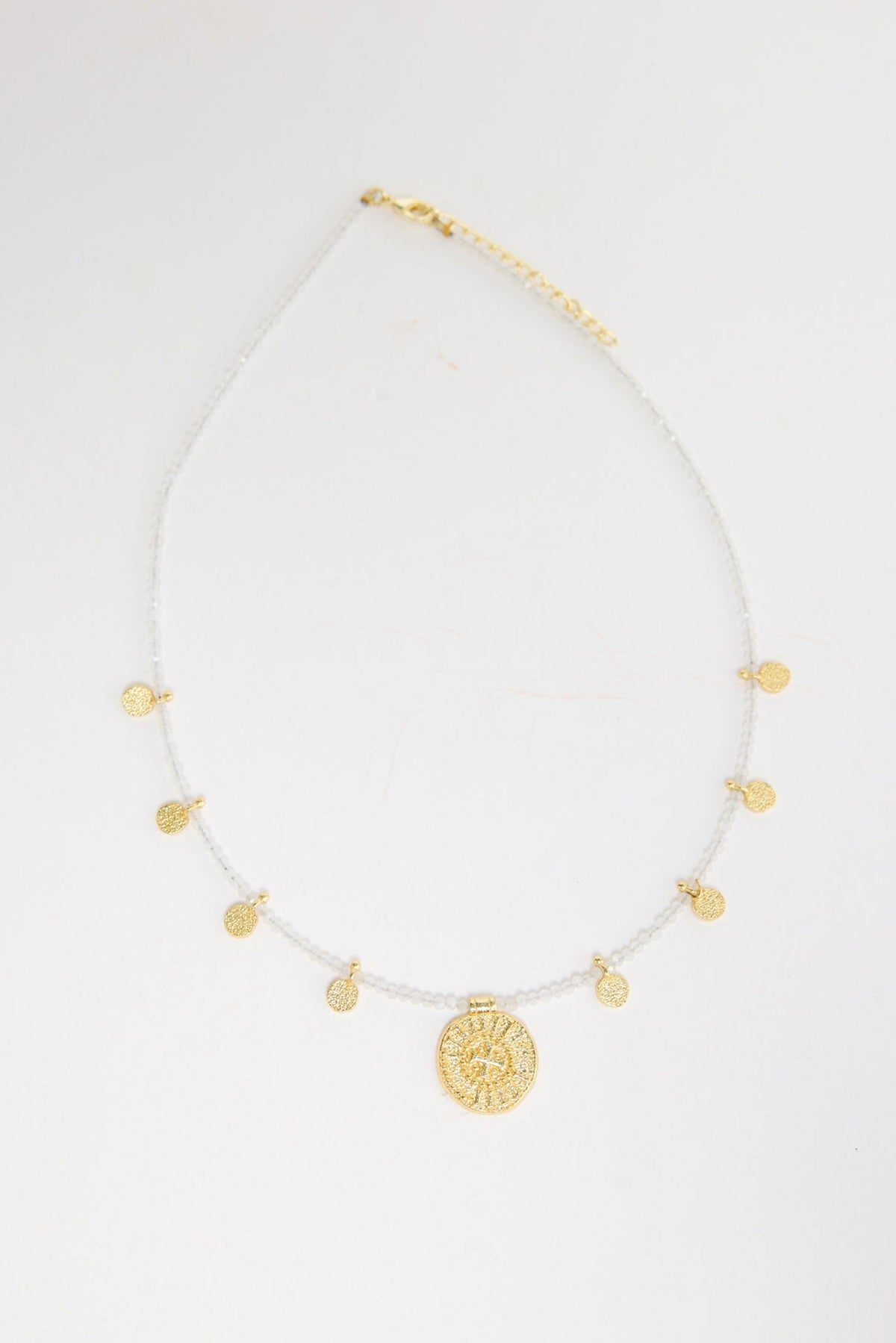 Delicate white crystal seed bead necklace with multiple small gold-tone coin charms and a larger engraved gold medallion pendant at the centre, shown flat on a white background