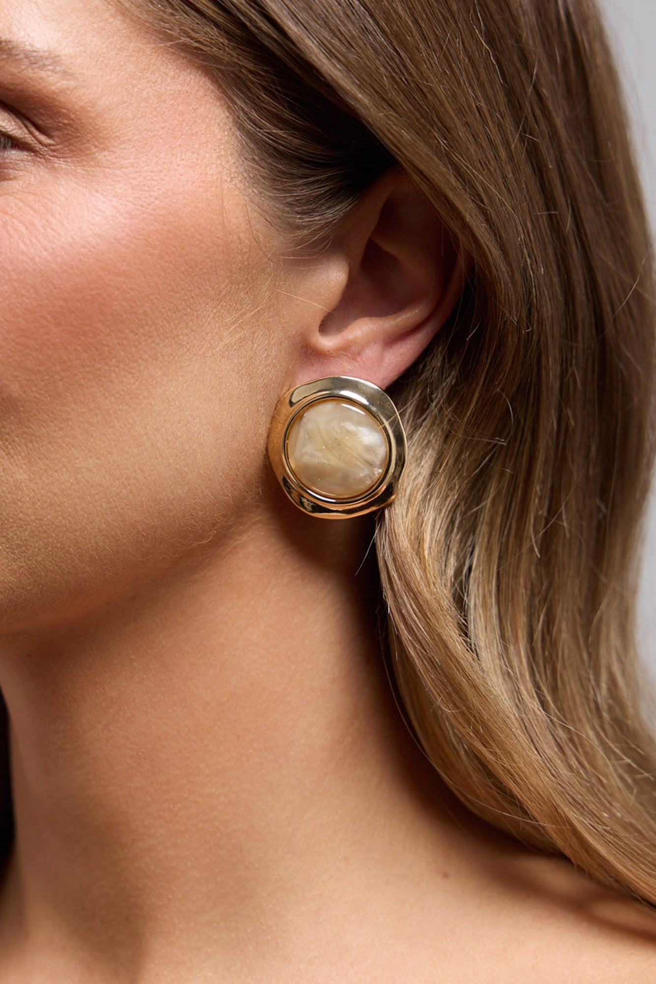 Rover Earrings in cream worn on the ear, showing the gold-tone frame and pearl-look resin centre up close