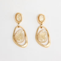 Pair of gold-tone drop earrings with a small oval cream resin stud and a large organic-shaped open gold hoop holding a cream resin stone, laid flat on a white background