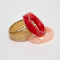 Set of three chunky resin bangles in cherry red, olive-gold and blush pink marbled finishes, stacked and interlocked together on a white background