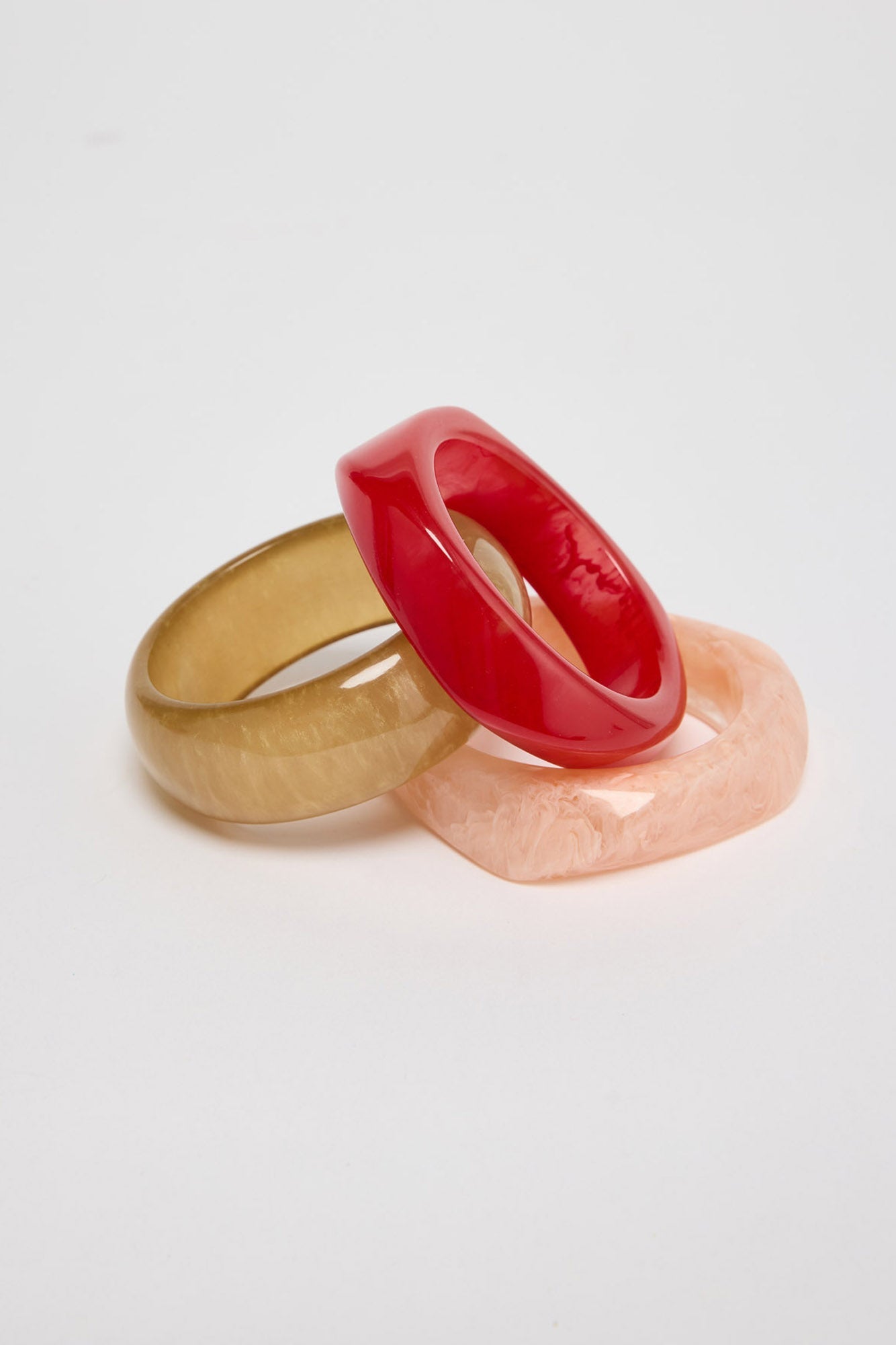Set of three chunky resin bangles in cherry red, olive-gold and blush pink marbled finishes, stacked and interlocked together on a white background