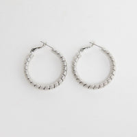 Neela Earrings in Silver – twisted, textured silver-tone hoop earrings shown flat on a white background.
