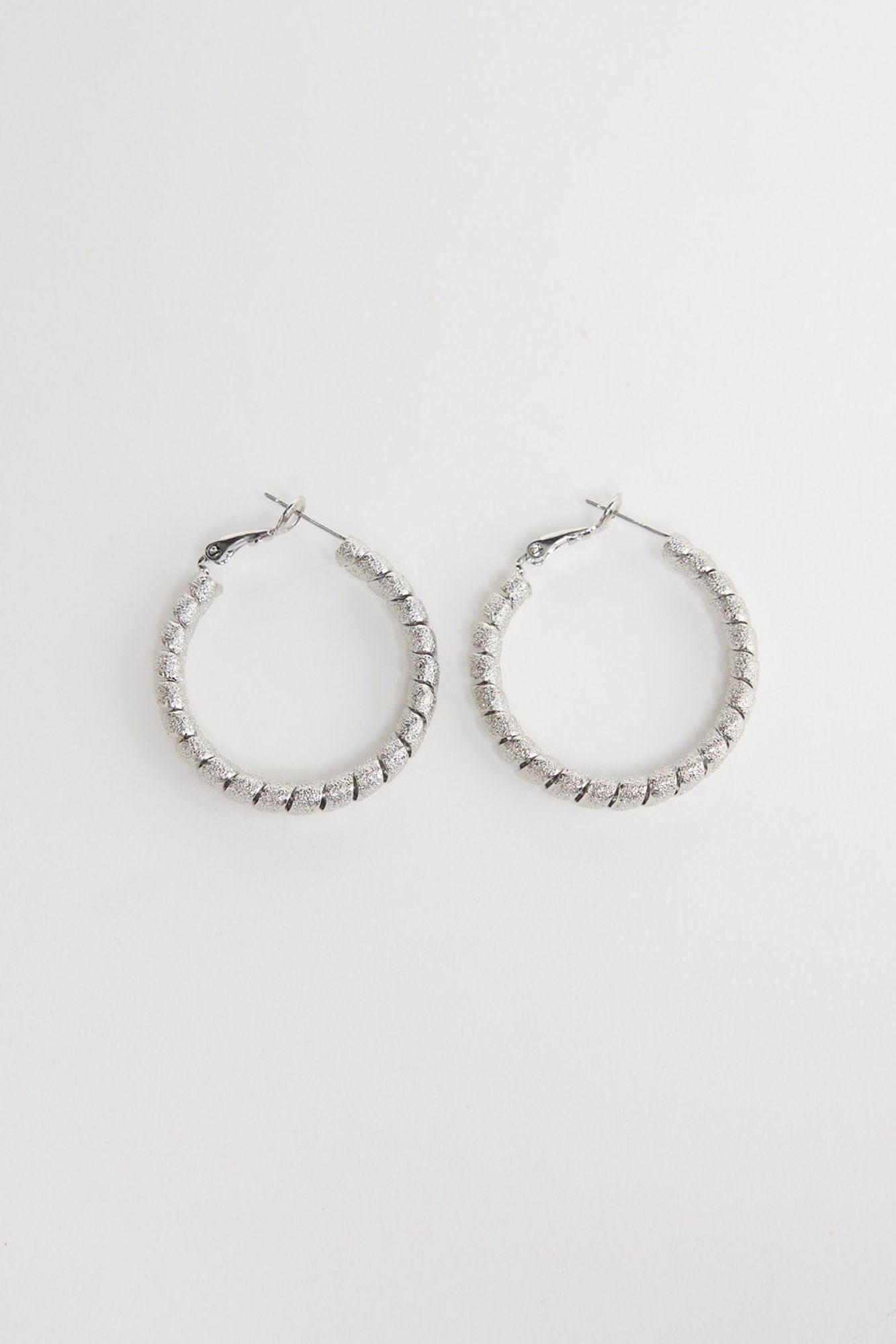 Neela Earrings in Silver – twisted, textured silver-tone hoop earrings shown flat on a white background.