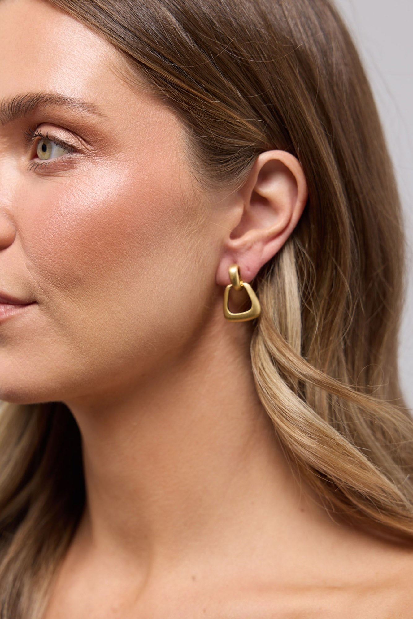Swell Earrings in Gold worn on the ear, showing the rounded-triangle geometric drop against the lobe.