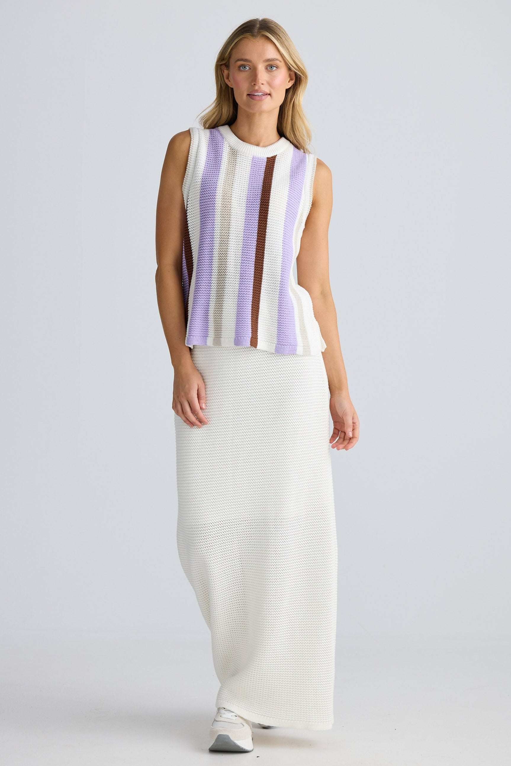 Woman wearing a sleeveless striped top and white pants on a light gray background