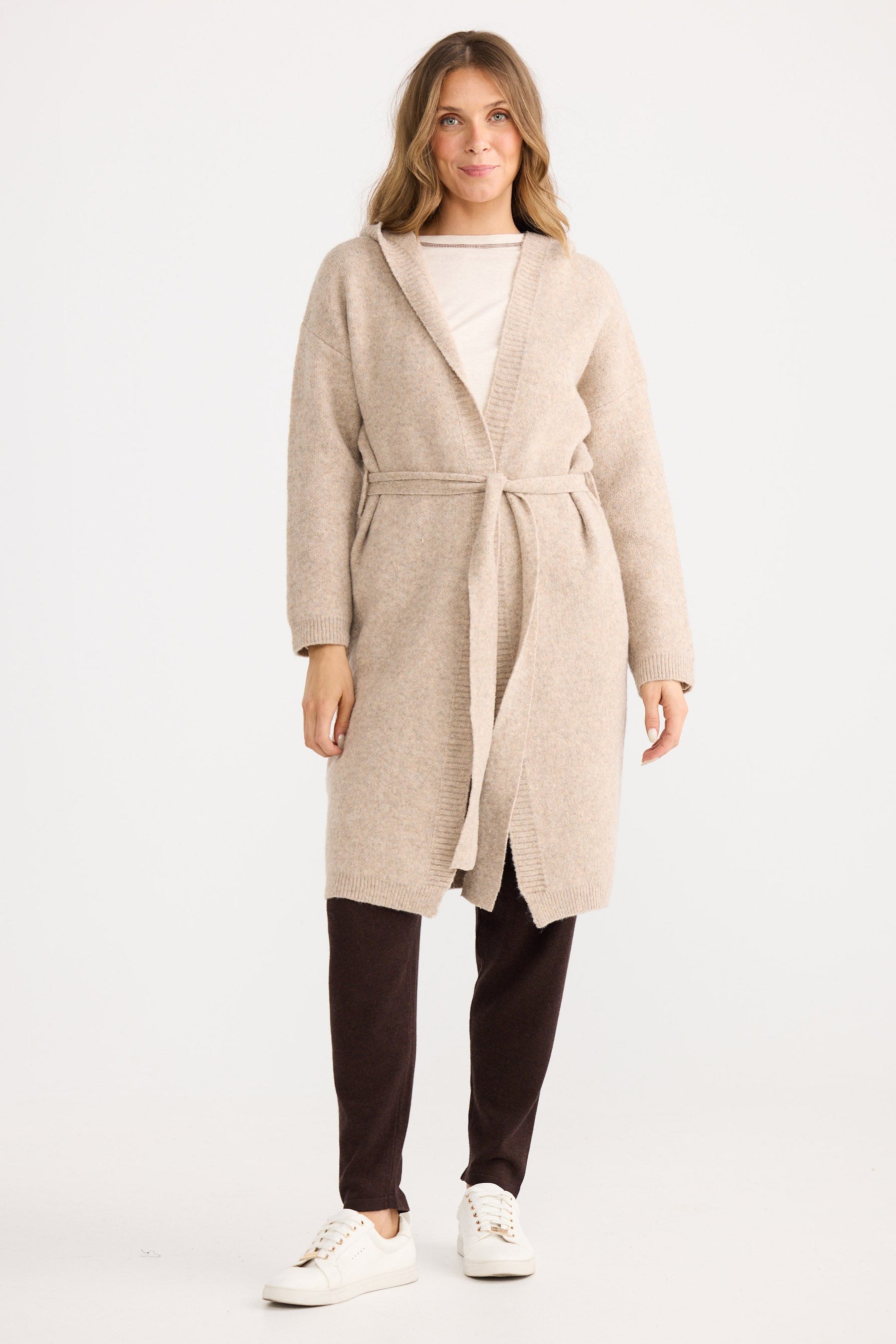 Woman wearing a beige cardigan over a white top and dark pants on a white background