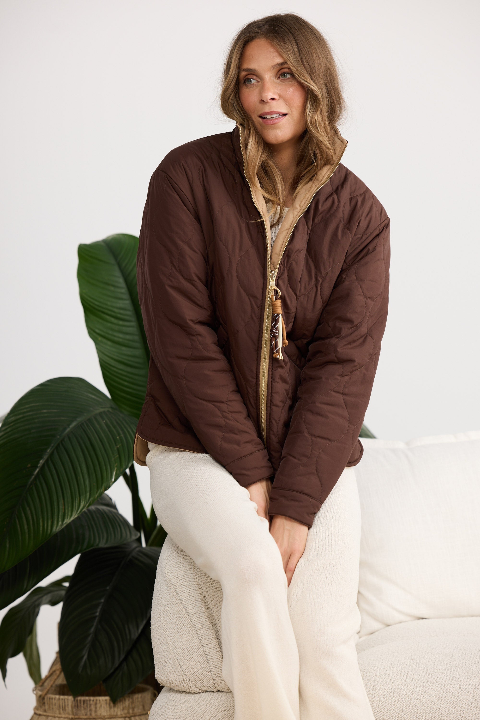 Woman wearing a brown quilted jacket sitting on a white couch with a plant in the background